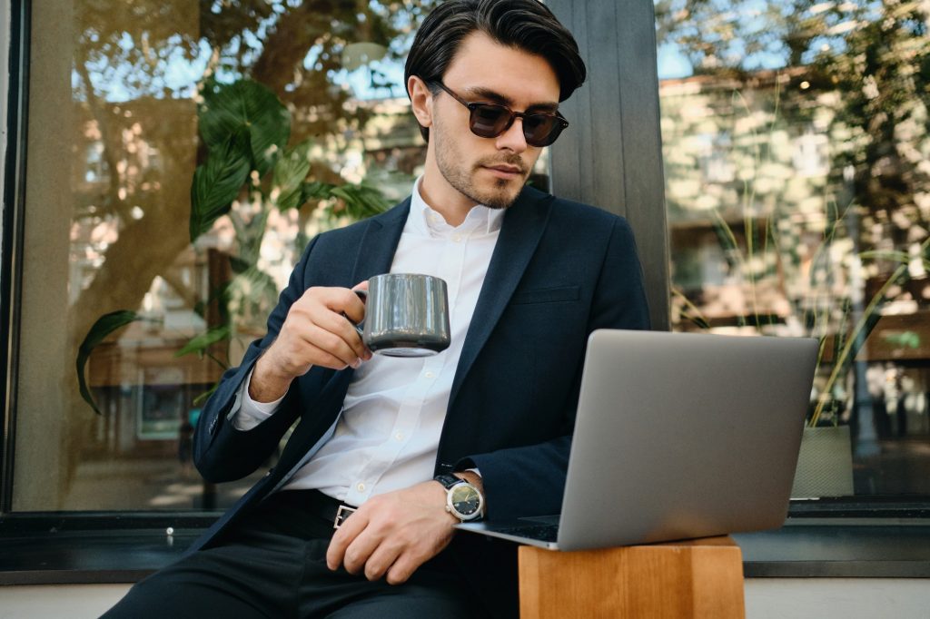 Young stylish man in jacket and sunglass drinking coffee dreamily working on laptop at street cafe